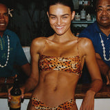Woman in leopard print bikini holding a beer at a bar with two men behind her.
