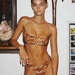 Woman wearing a leopard print bikini in an indoor setting with a shelf and decorative items.