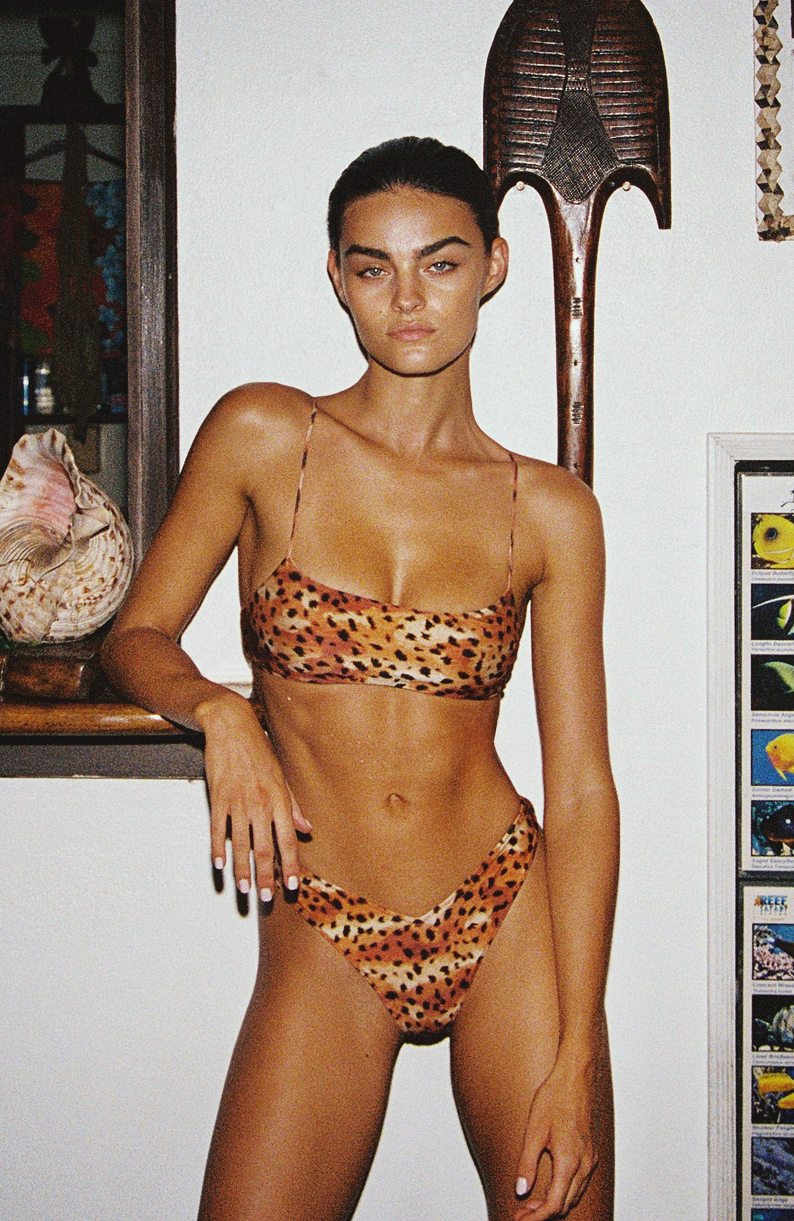 Woman wearing a leopard print bikini in an indoor setting with a shelf and decorative items.