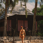 Person in a bikini standing on a sandy path leading to a thatched-roof hut with palm trees in the background.