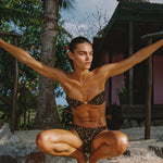 Person in a leopard print bikini posing outdoors with palm trees and a hut in the background