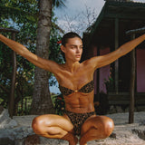 Person in a leopard print bikini posing outdoors with palm trees and a hut in the background