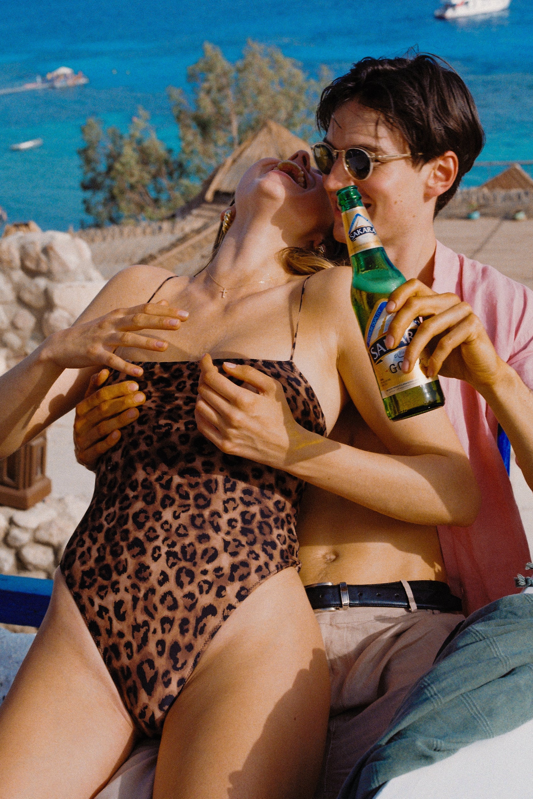 A person in a leopard print one-piece swimsuit is sitting close to another person who is holding a beverage bottle. They appear to be at a beach or poolside setting.
